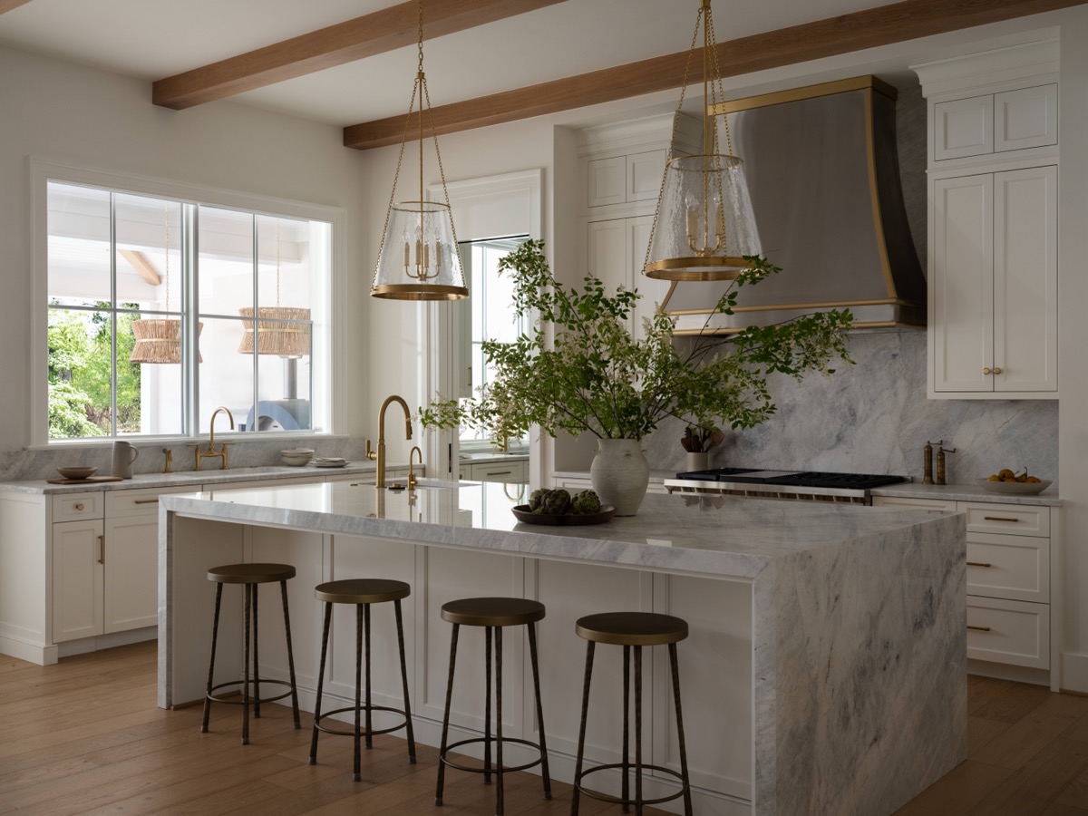 Bright white kitchen with marble island, brass fixtures, and four stools under a skylight-like window display.