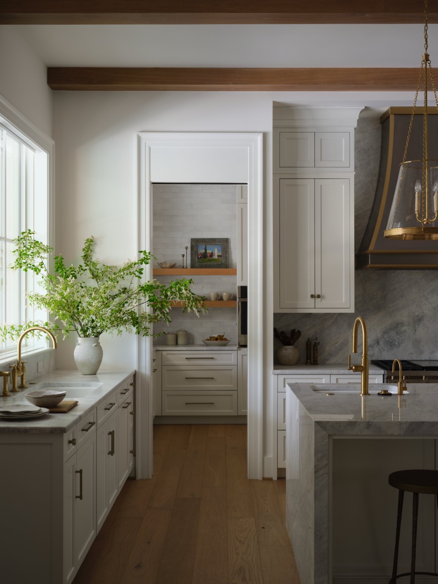 Bright, traditional white kitchen with marble island, brass fixtures, and a large vase of green branches by a window; open doorway reveals a pantry area with shelves and bowls.
