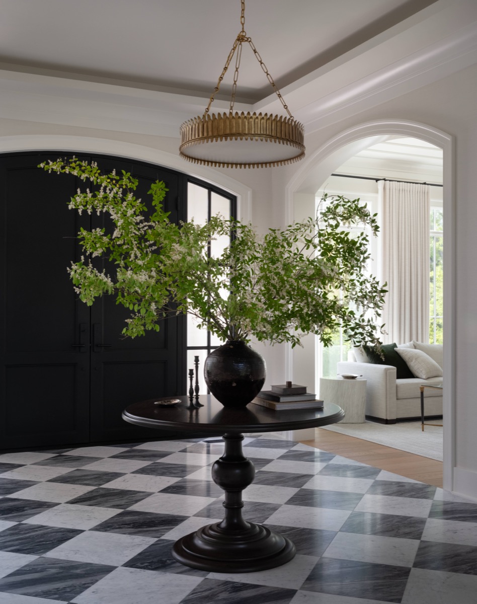 Dark wooden pedestal table with a large black vase of greenery in a foyer, a black armoire, and a black-and-white checkered floor with a gold chandelier overhead