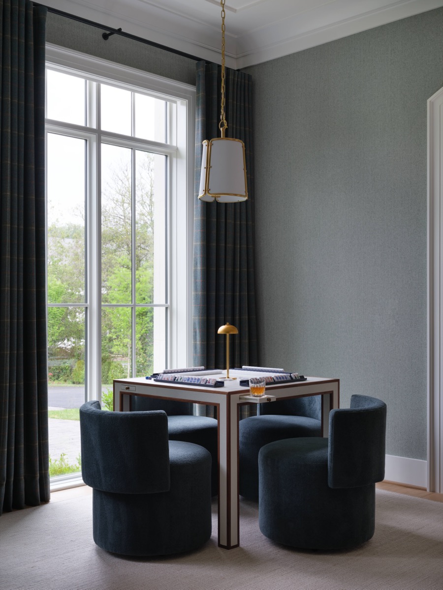 Four dark blue velvet chairs circle a square white table with gold accents, beneath a hanging pendant light by a large window with navy curtains.