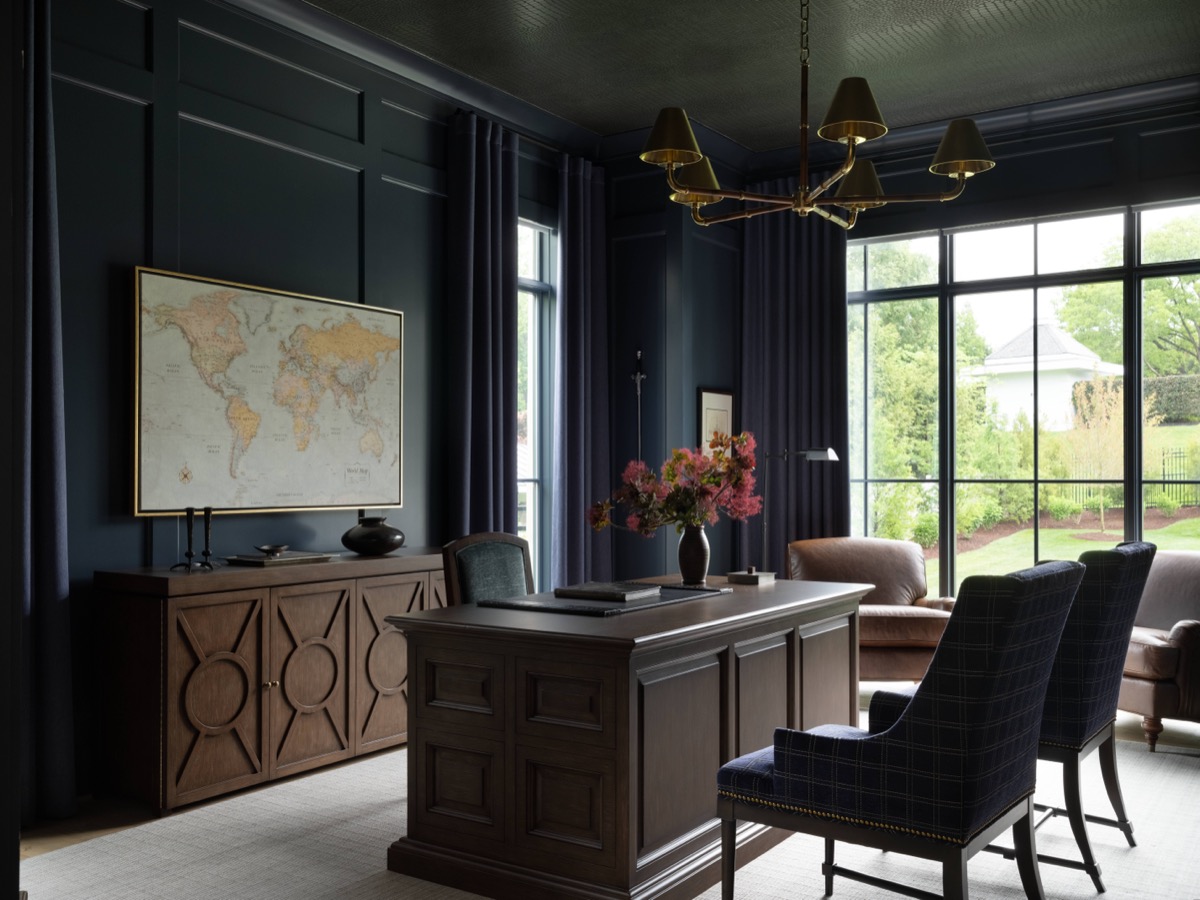 Dark wood executive office with navy walls, a world map on the sideboard, and a large desk facing two plaid chairs by tall windows.