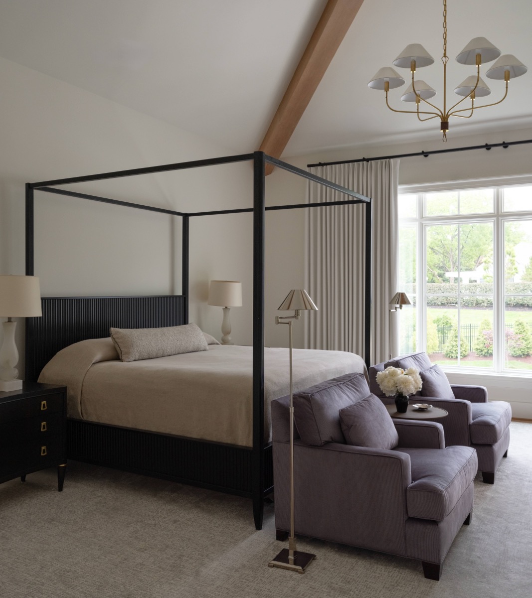 Dark metal four‑poster bed with beige bedding and ribbed headboard, flanked by nightstands with lamps in a bright bedroom. A purple seating area and large window with cream curtains nearby.